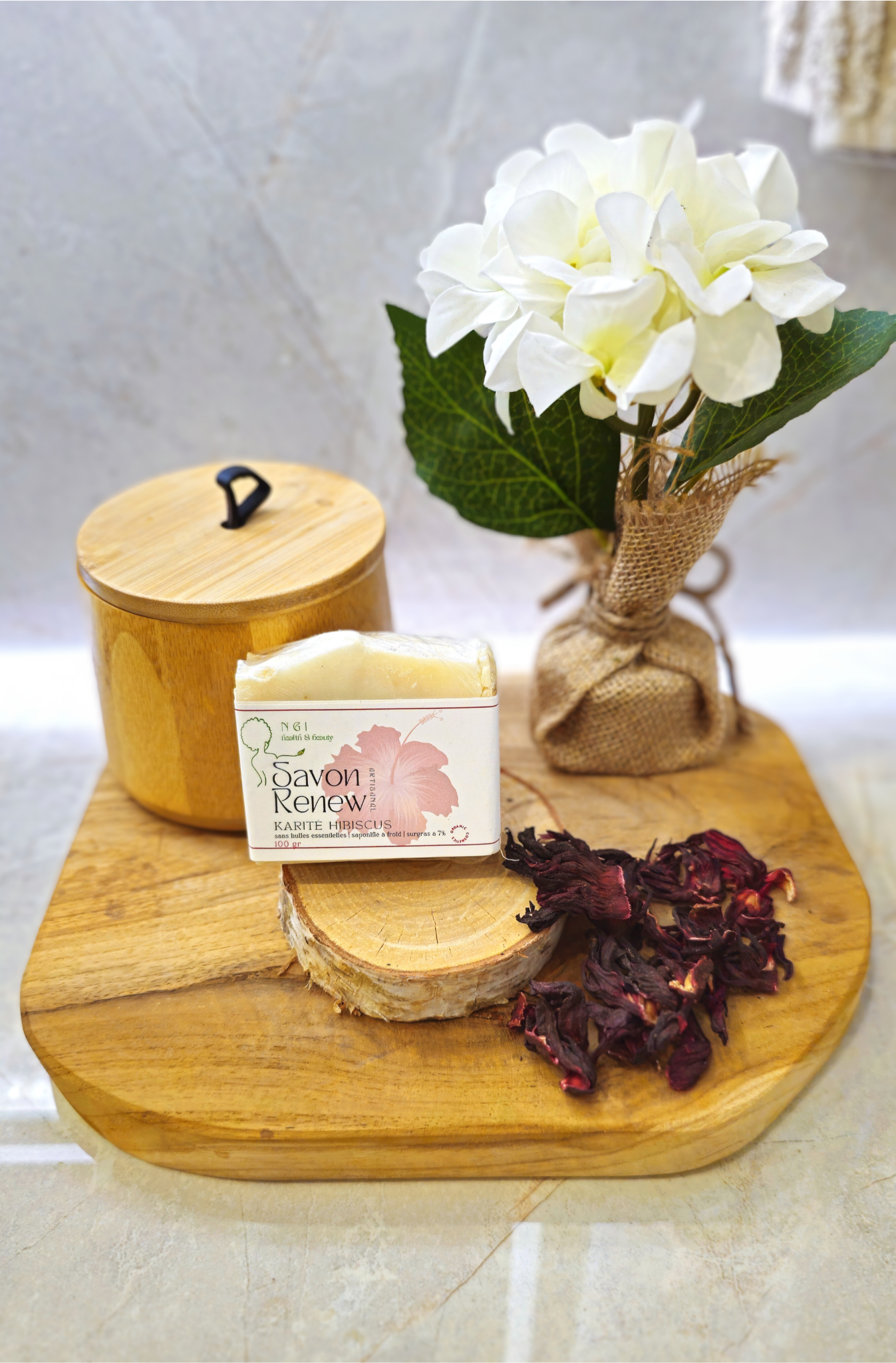Savon Karité/Hibiscus présenté sur un plateau en bois avec fleurs et pétales d’hibiscus, idéal pour un soin exfoliant et hydratant au quotidien.